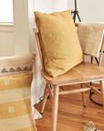 Nira Ochre Hand Spun Organic Cotton Handloom Pillow on a cozy wooden chair with textured fabric.