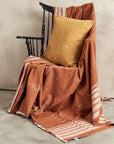 Nira Ochre Hand Spun Organic Cotton Handloom Pillow on a chair with woven throw blanket.