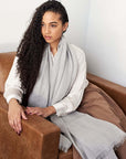 Woman wearing a Pebble Light Gray Hand Woven Soft Linen Scarf, sitting comfortably on a brown couch.
