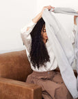 Woman styling Pebble Light Gray Hand Woven Soft Linen Scarf while sitting on a couch.