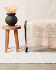 Butterscotch White Handloom Textured Plush Merino Throw displayed on a bench beside a wooden stool and vase with decorative plant.