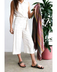 Woman in a light outfit holding Barque Clay Modern Artisan Striped Soft Merino Handwoven Scarf next to a plant.