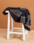Walnut Black Yak With Hand Knotted Merino Stripes Handloom Throw draped on a white stool against a warm background.