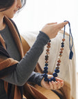 Woman holding and looking at  Mihika silk beaded necklace, with a brown cashmere scarf draped over her. 