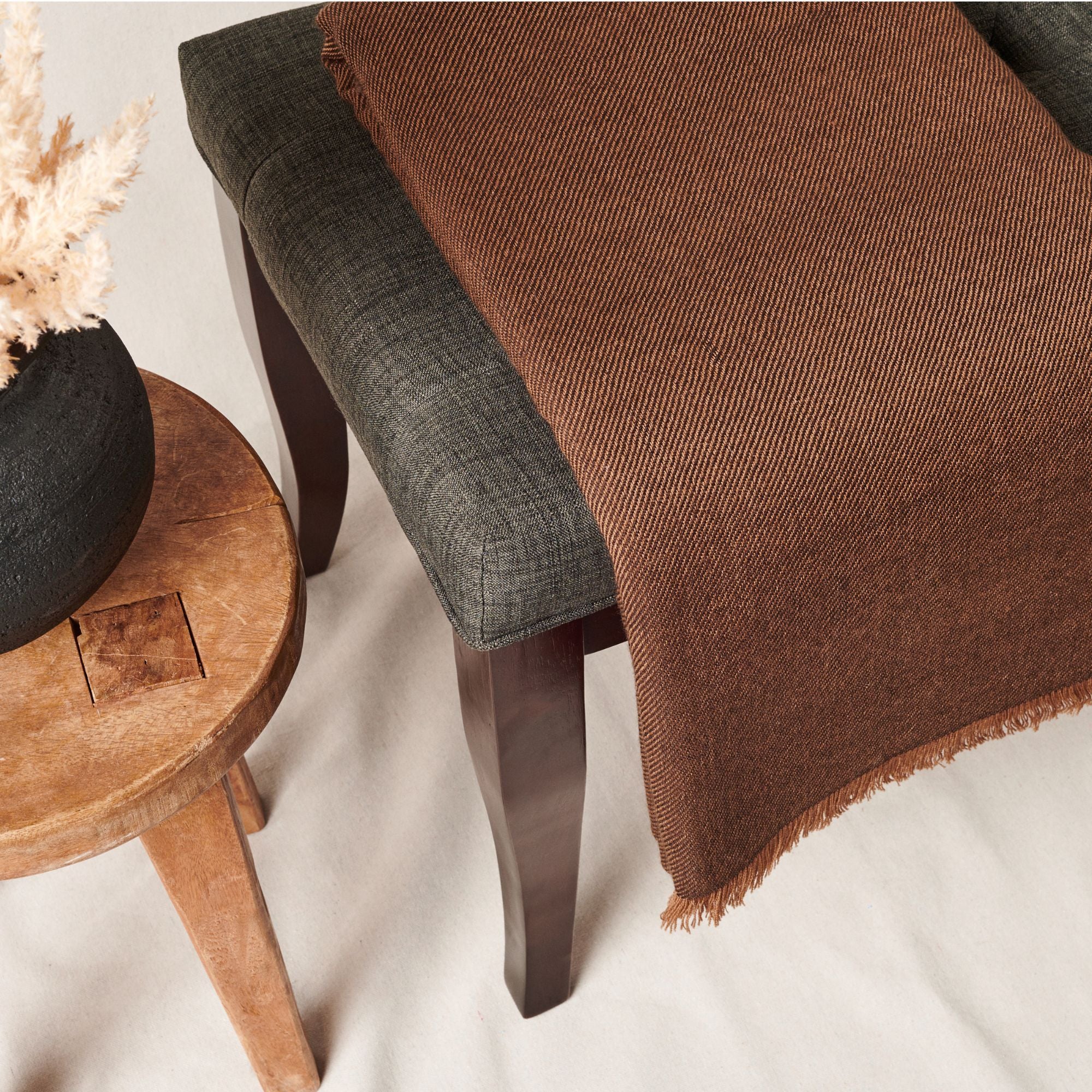 Hay Brown Twill Merino Handwoven Throw draped on a dark gray chair beside a wooden stool with dried plants.