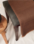 Hay Brown Twill Merino Handwoven Throw draped on a dark gray chair beside a wooden stool with dried plants.