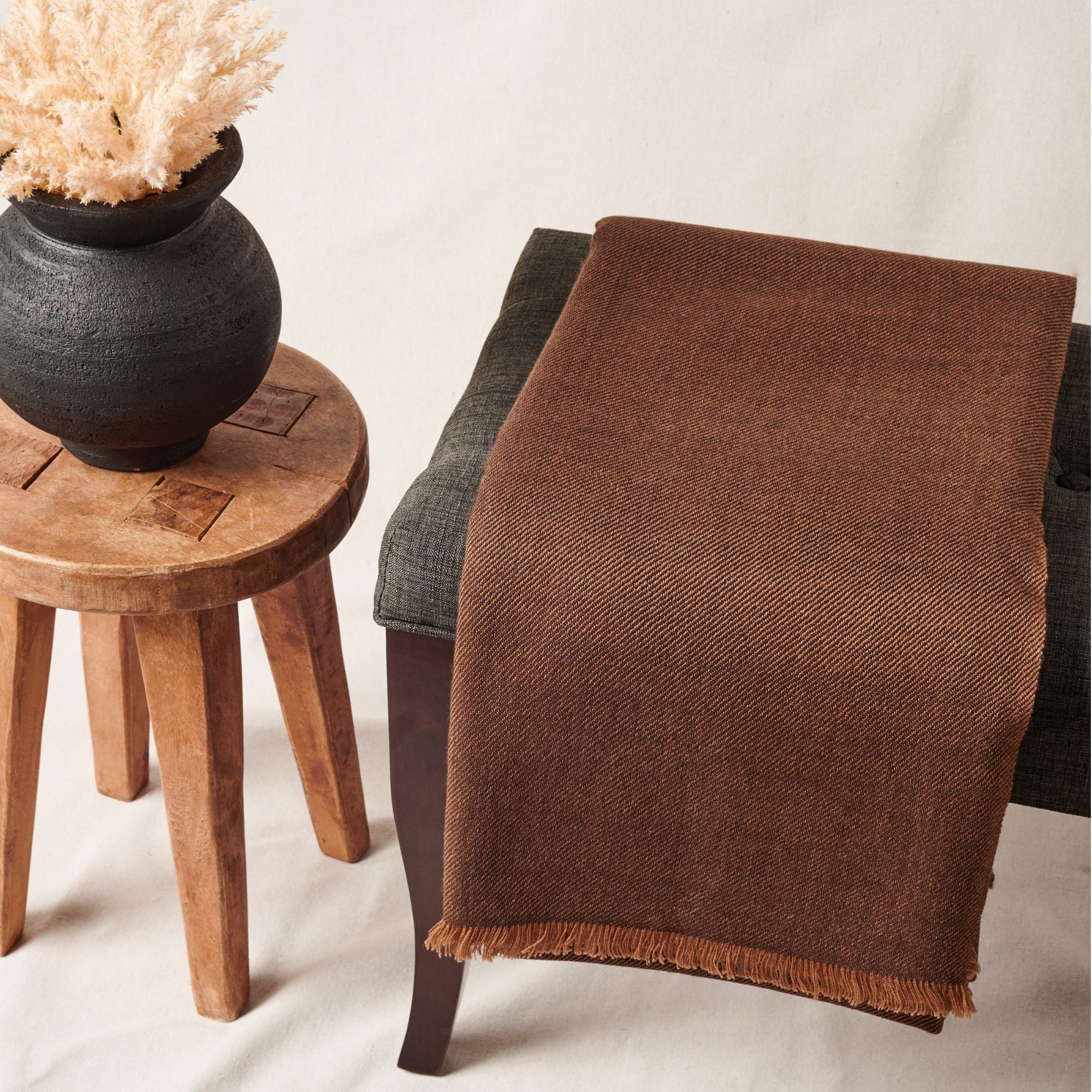 Hay Brown Twill Merino Handwoven Throw draped over a chair beside a vase, showcasing its soft brown texture and fringe.