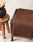 Hay Brown Twill Merino Handwoven Throw draped over a chair beside a vase, showcasing its soft brown texture and fringe.
