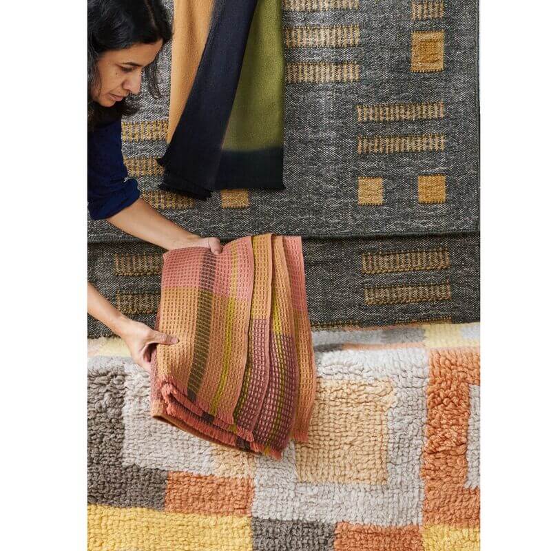 Person holding a soft, textured rug in shades of pink and orange next to various geometric pattern rugs.