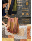 Person holding a soft, textured rug in shades of pink and orange next to various geometric pattern rugs.