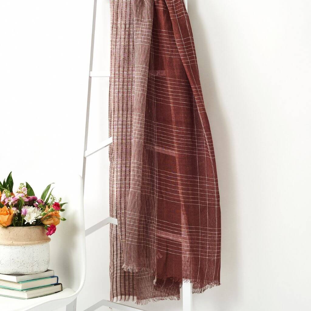 Auro Cinnabar Handwoven Earthy Rust Maroon Linen Scarf displayed on a ladder next to flowers.