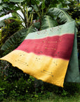 Avocado Green Multi Color Warm Hues Hand Woven Hand Knotted Textured Merino Throw displayed outdoors among lush greenery.