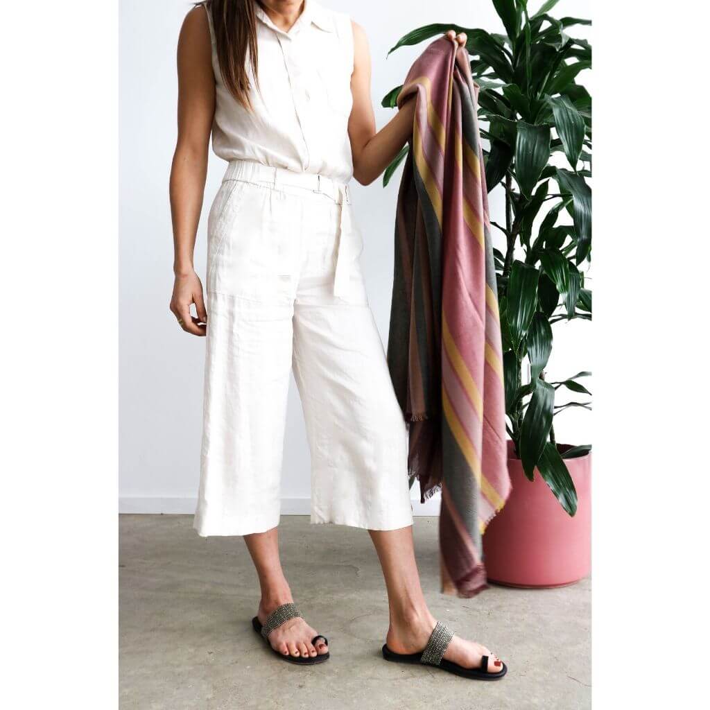 Woman in a light outfit holding Barque Clay Modern Artisan Striped Soft Merino Handwoven Scarf next to a plant.
