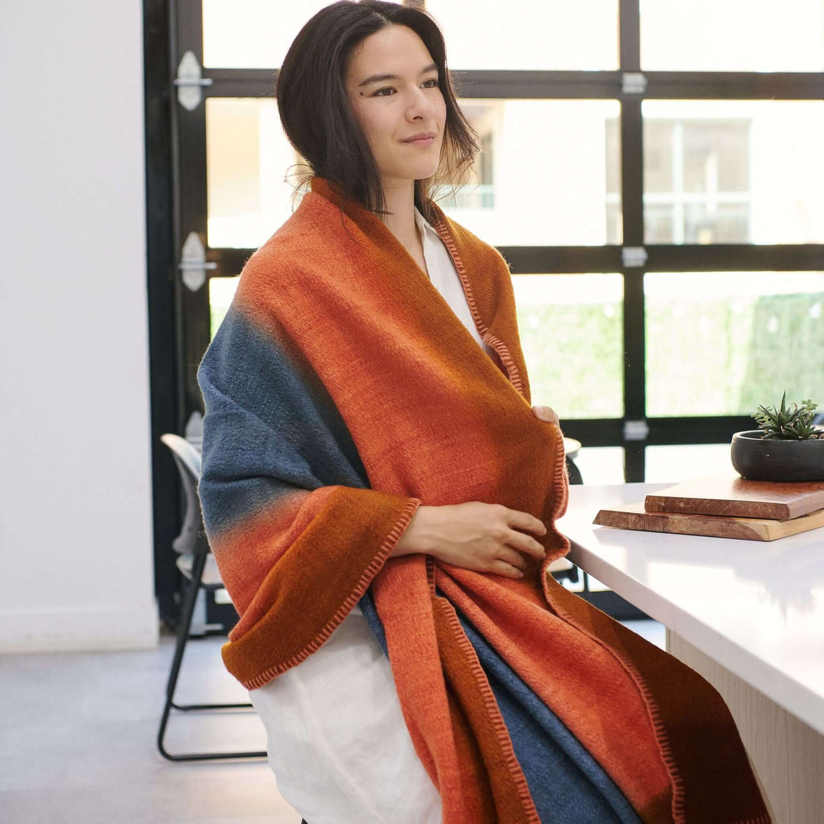 Ember Apricot In Orange and Blue Shades Handwoven Merino Throw Blanket draped over a woman in a modern setting.