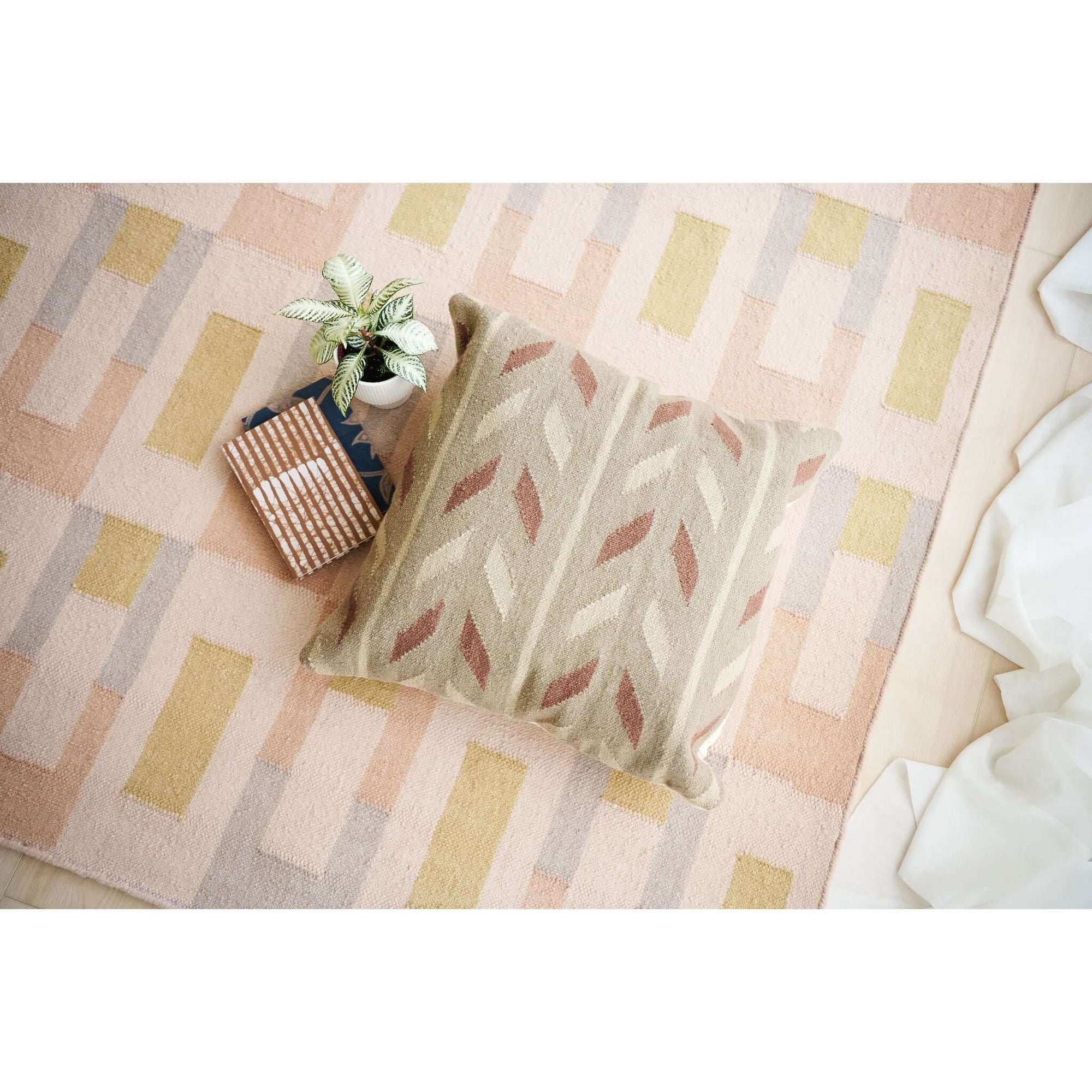 Ero Green Brown Geometric Pattern Hand Woven Large Pillow displayed on a textured area rug with a plant and books.