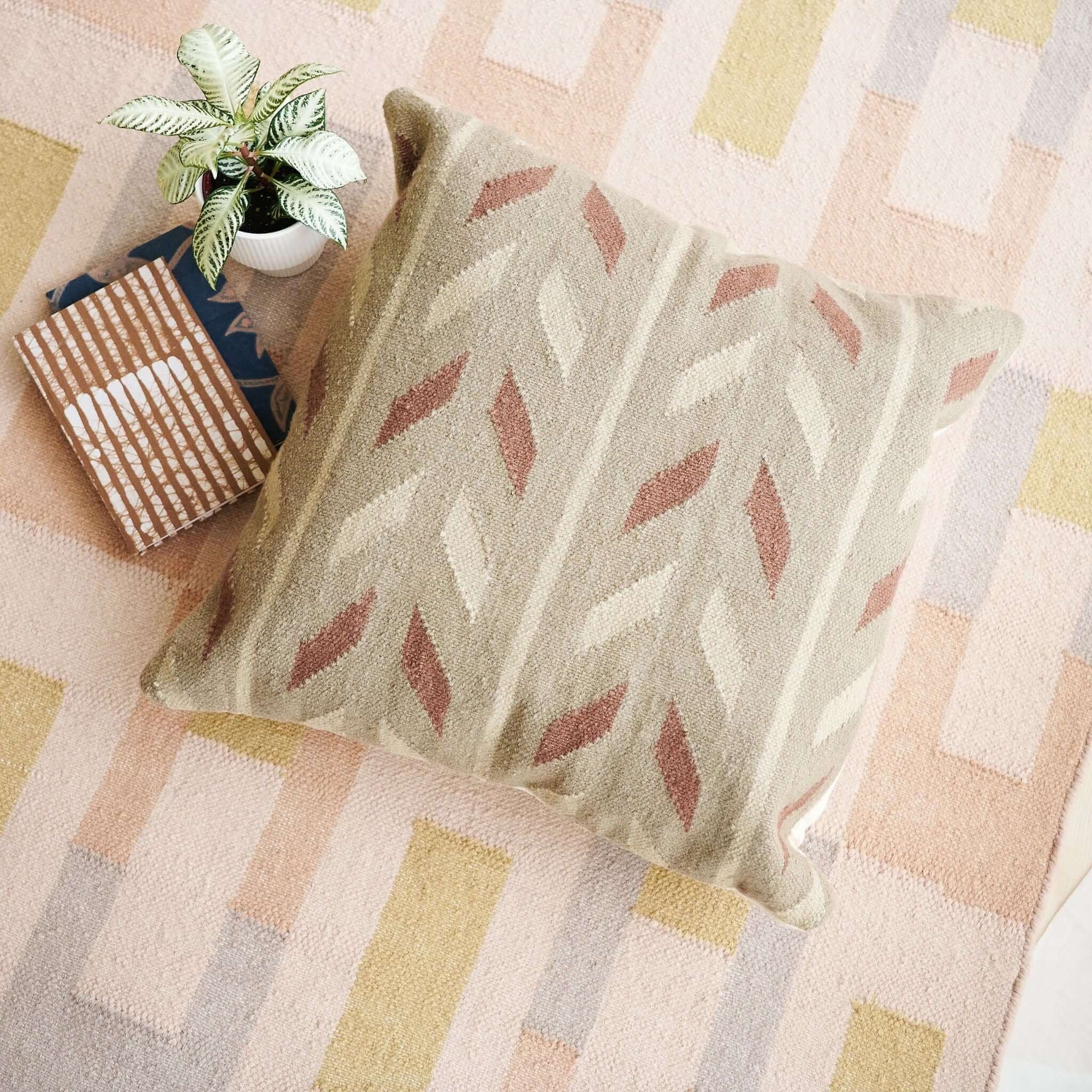 Ero Green Brown Geometric Pattern Hand Woven Large Pillow on a patterned rug with a plant and books.
