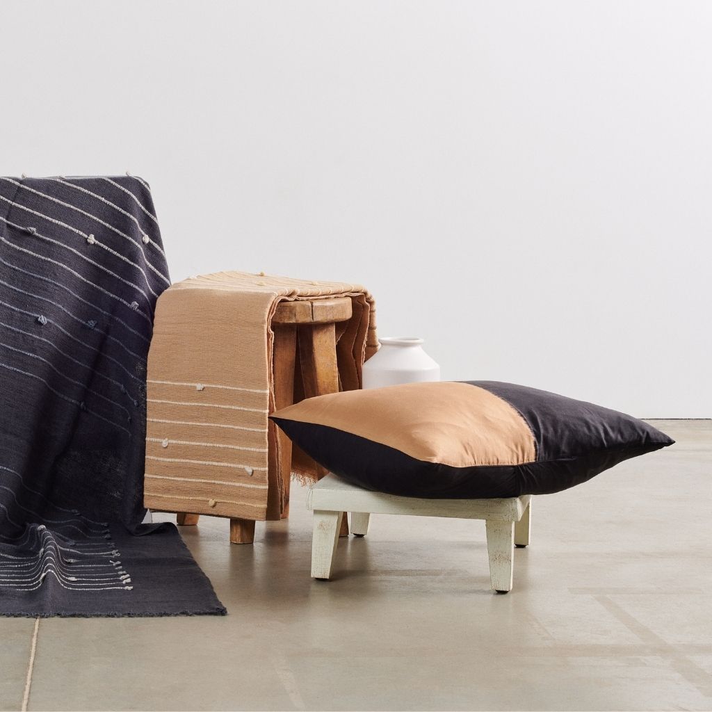 Kala Black & Gold Color Block Mulberry Silk Square Pillow on a wooden stool with textured blankets in a modern decor setting.