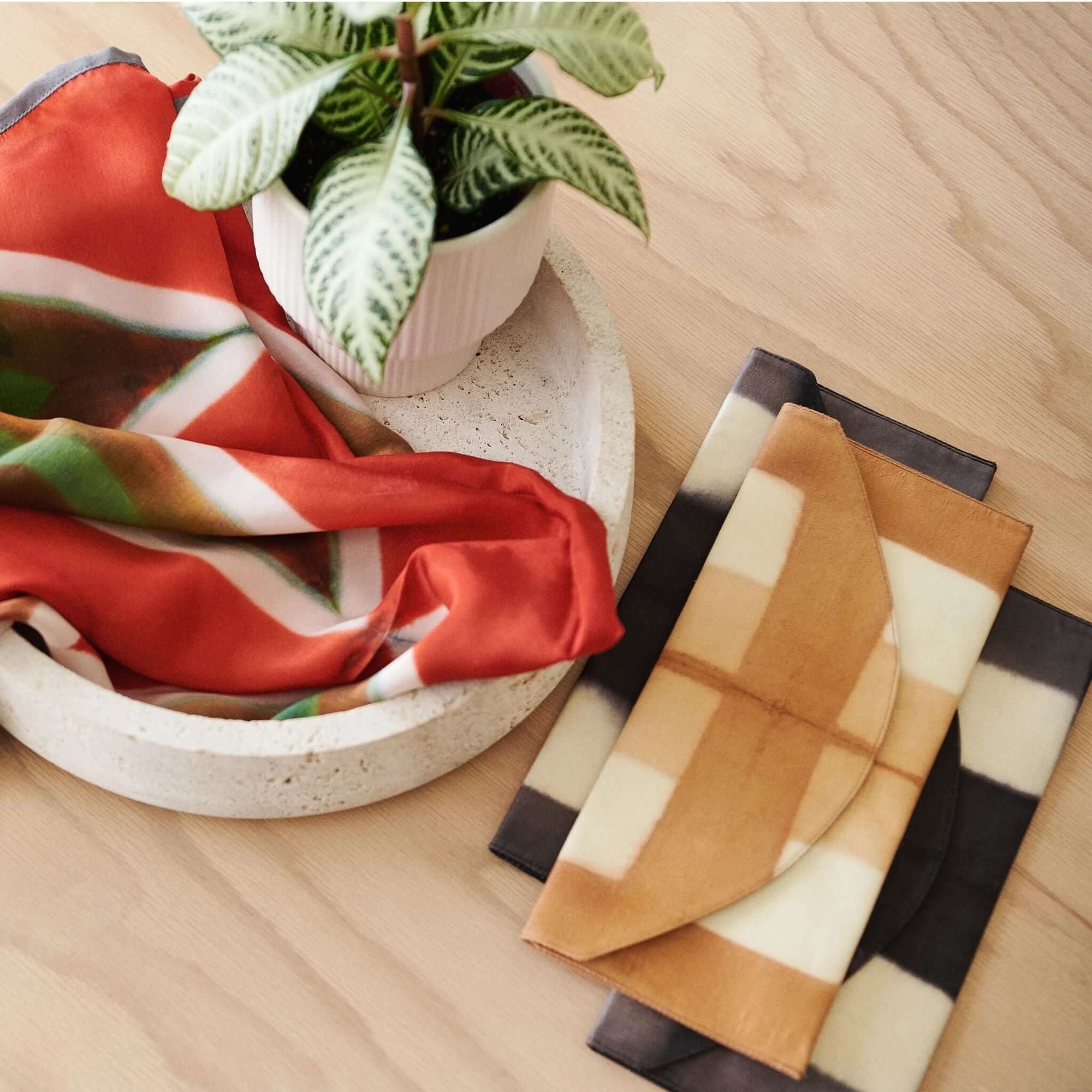 Melo Gold Mulberry Silk Shibori Hand Crafted Envelope Pouch displayed with a plant in a bowl and colorful silk scarves.
