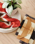 Melo Gold Mulberry Silk Shibori Hand Crafted Envelope Pouch displayed with a plant in a bowl and colorful silk scarves.