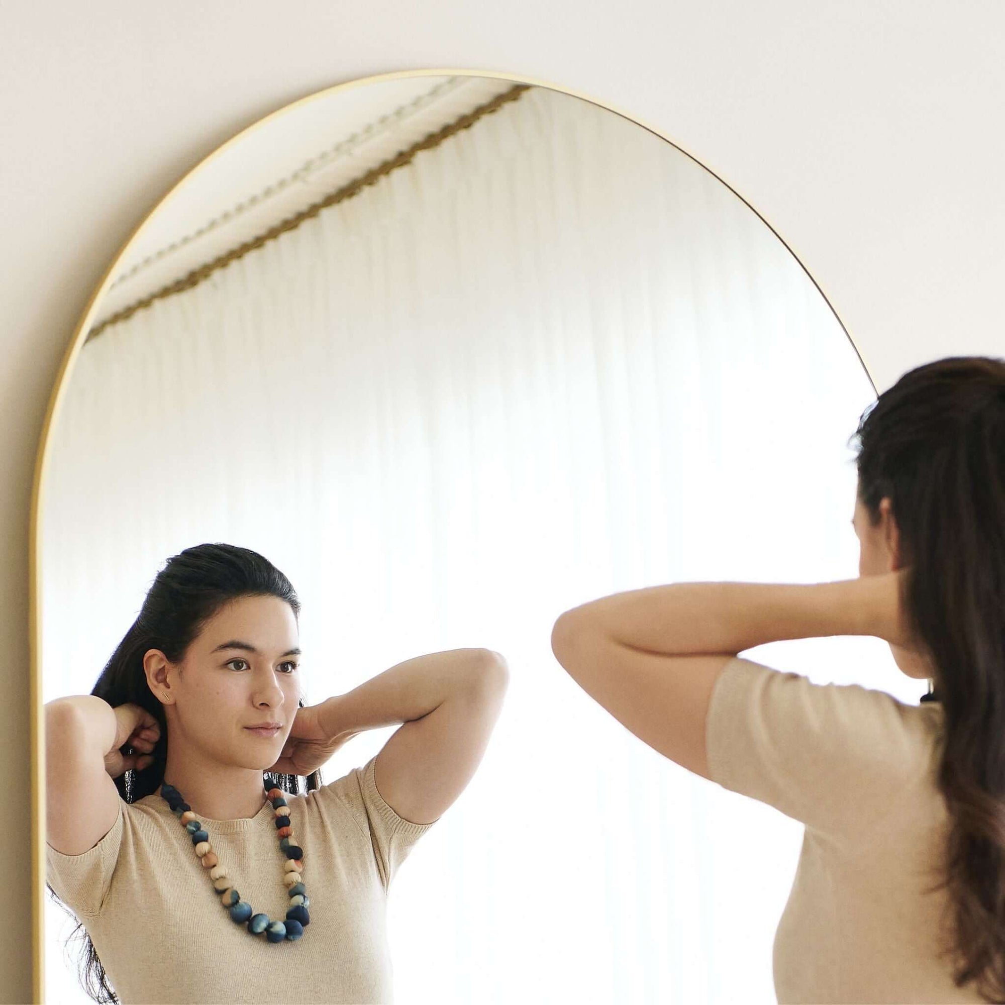 Woman wearing Mihika silk beaded necklace, adjusting her hair in front of a mirror, showcasing its beauty.