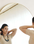 Woman wearing Mihika silk beaded necklace, adjusting her hair in front of a mirror, showcasing its beauty.